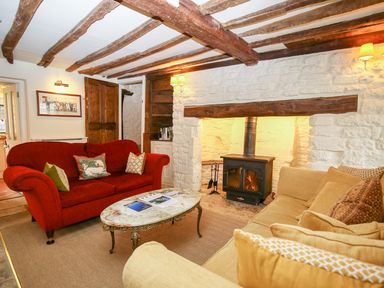 A living room with a red sofa a beige sofa a stone fireplace with a wood stove and wooden ceiling beams at Murton Cottage in Burford