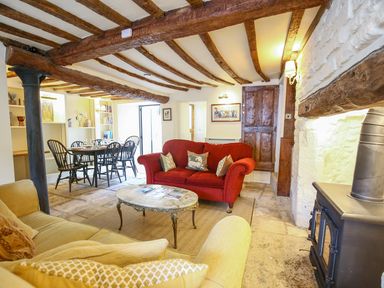 A living room with a red sofa a beige sofa a coffee table a dining table with chairs wooden beams on the ceiling and a stove at Murton Cottage in Burford