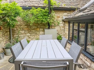 An outdoor patio with a rectangular table and six chairs next to stone walls and windows at Murton Cottage in Burford