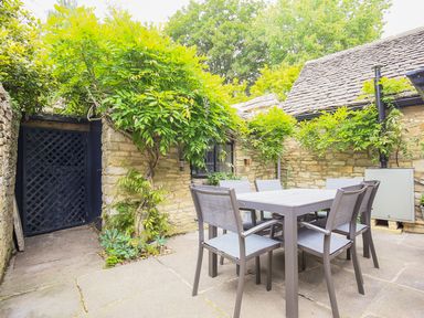 An outdoor patio area with a table and six chairs surrounded by stone walls and greenery at Murton Cottage in Burford