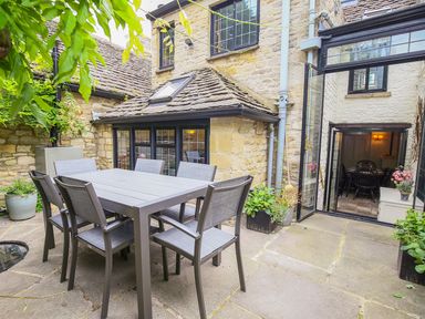 An outdoor patio with a dining table and six chairs next to a stone building with windows and an open glass door at Murton Cottage in Burford
