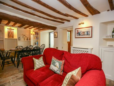 A living room with a red sofa and pillows a dining table with chairs and wooden ceiling beams at Murton Cottage in Burford