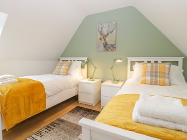 A bedroom with two single beds with yellow throws and plaid pillows two white nightstands with green lamps and a deer painting on a green wall at The Forge in Shipham