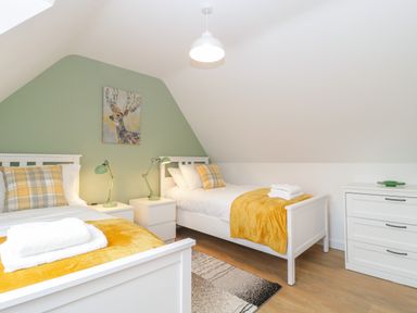 A bedroom with two white beds with yellow blankets and plaid pillows a green accent wall with deer artwork and white furniture at The Forge in Shipham