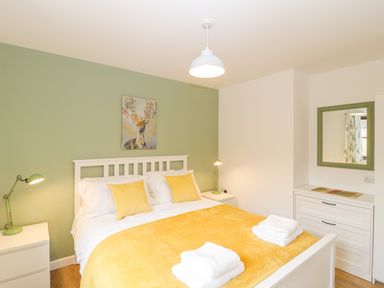 A bedroom with a white bed with yellow pillows and blanket two bedside tables with lamps and a mirror above a dresser at The Forge in Shipham