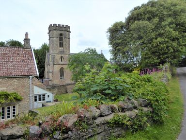 A stone church and a house next to a stone wall with plants and flowers along a path at The Forge in Shipham