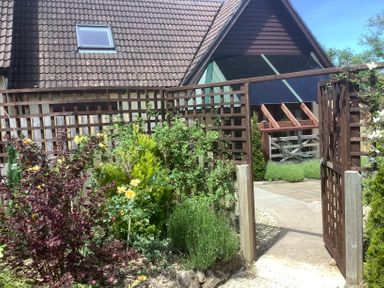 A garden with plants and flowers behind a wooden trellis gate and a house with a tiled roof at The Forge in Shipham