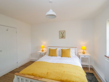A bedroom with a double bed yellow pillows and a yellow blanket two white bedside tables with yellow lamps and a framed picture on the wall at Mendip Cottage in Shipham