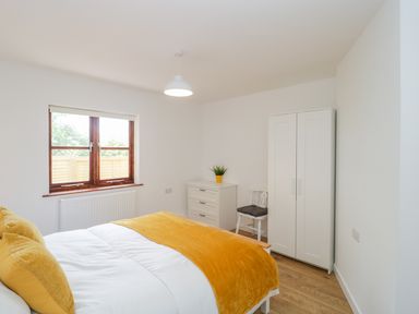 A bedroom with a bed with yellow pillows and throw a white dresser with a plant a white chair and a white wardrobe at Mendip Cottage in Shipham