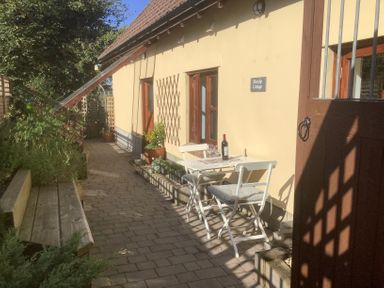 A patio with a small white table and two chairs against a yellow cottage wall with plants and a wooden bench at Mendip Cottage in Shipham
