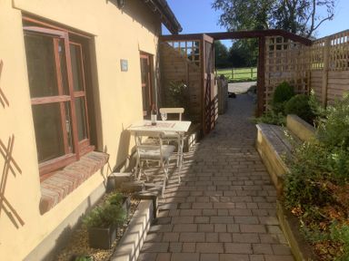 A small outdoor seating area with a table and two chairs beside a yellow building and a pathway with garden beds and wooden fencing at Mendip Cottage in Shipham