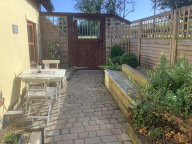 A small outdoor patio area with a white table and chairs a bottle of wine and glass wooden gate and raised garden beds at Mendip Cottage in Shipham