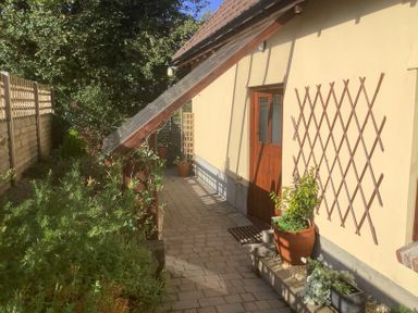 An outdoor paved walkway with potted plants alongside a yellow house with a wooden door and wooden lattice in Shipham