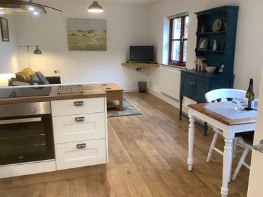 A living area with a kitchen island stove white cabinets wooden floor a blue cabinet a dining table with chairs and a small television on a wall shelf at Mendip Cottage in Shipham