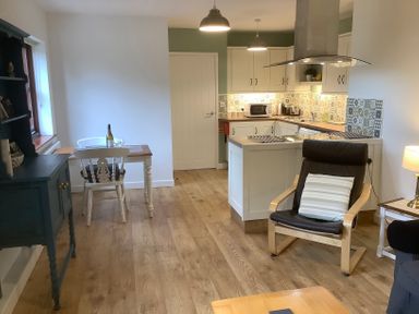 A kitchen and dining area with wooden floor a black armchair and a small dining table with two chairs at Mendip Cottage in Shipham