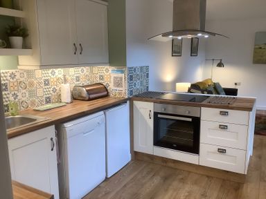 A kitchen with white cabinets wooden countertops a built-in oven a stovetop a toaster a dishwasher and a patterned tile backsplash at Mendip Cottage in Shipham