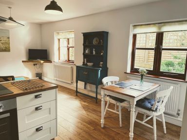 A kitchen and dining area with a wooden table, two white chairs, a blue cabinet, a wall-mounted TV, and wooden flooring at Mendip Cottage in Shipham