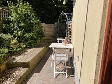A small outdoor patio with a white table and two chairs a bottle and glasses on the table and green plants along a wooden fence at Mendip Cottage in Shipham
