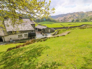 A stone farmhouse with outbuildings and green fields with hills in the background at Tilberthwaite Farm Cottage in Coniston