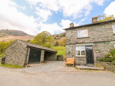 A stone house with a black door and windows next to a stone garage with black doors and a bench outside at Tilberthwaite Farm Cottage in Coniston
