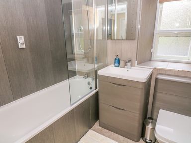A bathroom with a bathtub shower, sink with cabinet, window, toilet, and small trash bin at Tilberthwaite Farm Cottage in Coniston