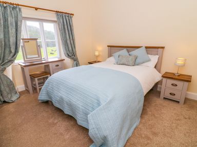 A bedroom with a bed blue bedding two bedside tables with lamps a dressing table and window with curtains at Tilberthwaite Farm Cottage in Coniston