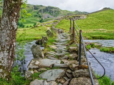 A stone bridge with metal railings over a river surrounded by green hills at Tilberthwaite Farm Cottage in Coniston
