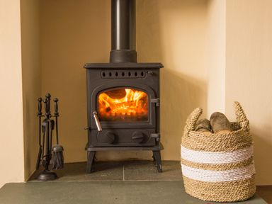 A wood burning stove with fire inside and a basket of firewood next to it at Tilberthwaite Farm Cottage in Coniston