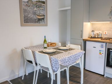 A dining area with a table set for two including wine and plates next to a kitchen space with cabinets and a refrigerator at Cuckoo Cottage in Bridport
