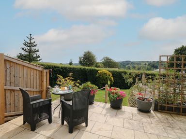A patio with two black chairs and a glass table with cups and plants overlooking a garden at Cuckoo Cottage in Bridport