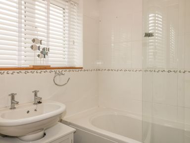 A bathroom with a sink and taps a bathtub with a glass shower screen and decorative fish on a windowsill at Dune Haven in Goonhavern near Perranporth