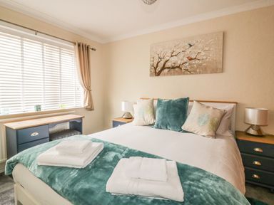 A bedroom with a bed and pillows a desk by the window and bedside tables with lamps at Dune Haven in Goonhavern near Perranporth