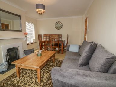 A living room with a gray sofa wooden coffee table dining table with chairs fireplace and wall clock at Dune Haven in Goonhavern near Perranporth
