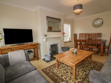 A living room with a wooden coffee table on a patterned rug a grey sofa a wooden dining table with chairs a fireplace and a wall-mounted mirror at Dune Haven in Goonhavern near Perranporth