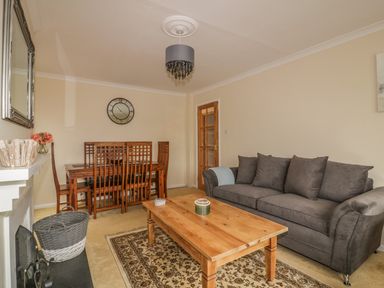 A living room with a gray sofa a wooden coffee table a dining table with chairs a wall clock and a fireplace at Dune Haven in Goonhavern near Perranporth