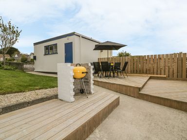 A backyard patio with a yellow barbecue grill a black outdoor table with chairs and an umbrella and a small building with a blue door at Dune Haven in Goonhavern near Perranporth