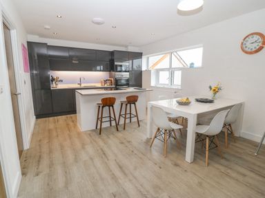 A kitchen with grey cabinets a white island with stools and a white dining table with chairs and flowers at Cysgod Y Capel in Benllech