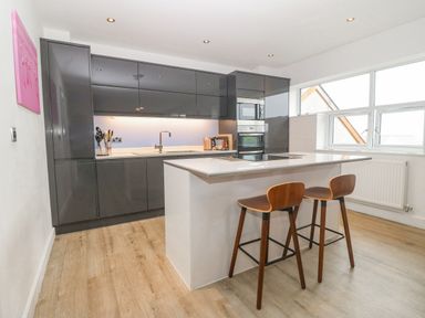 A modern kitchen with gray cabinets a white island with cooktop and two wooden stools at Cysgod Y Capel in Benllech