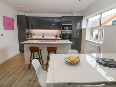 A kitchen with dark cabinets an island with two wooden stools and a dining table with a bowl of bananas at Cysgod Y Capel in Benllech