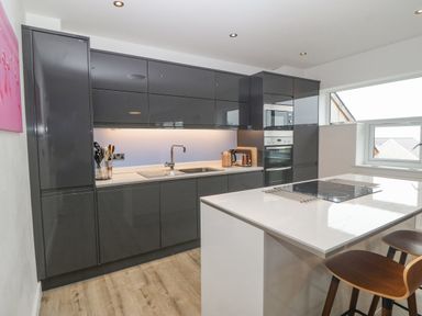 A kitchen with dark cabinets white countertops an island with a stovetop stools and a window at Cysgod Y Capel in Benllech