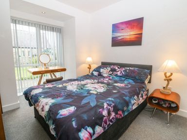 A bedroom with a double bed with floral bedding two bedside tables with lamps a wooden dressing table and vertical blinds on a window at Cysgod Y Capel in Benllech