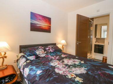 A bedroom with a floral bedspread a wooden nightstand with a lamp and an open door showing a bathroom at Cysgod Y Capel in Benllech