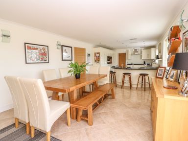 A dining area with a wooden table, white chairs, and a bench next to a kitchen with bar stools at Cadwgan in Aberporth