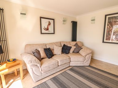 A beige corner sofa with cushions in a living room with framed pictures on the walls and a wooden side table at Cadwgan in Aberporth