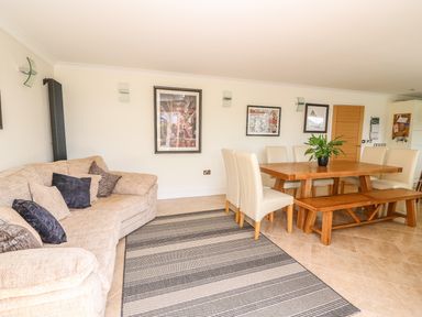 A living room with a beige sofa and cushions a wooden dining table with chairs and a bench a plant on the table and framed pictures on the wall at Cadwgan in Aberporth