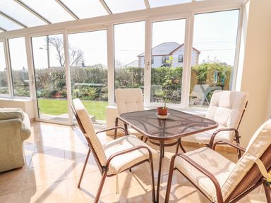 A sunroom with four cushioned chairs around a metal table with a potted plant and glass windows showing a garden at Cadwgan in Aberporth