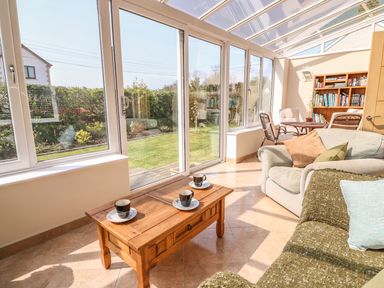 A sunroom with large windows wooden coffee table with cups sofas a dining table with chairs and a bookshelf at Cadwgan in Aberporth