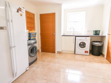 A laundry room with a refrigerator washing machines a countertop with a sink wooden doors and a trash bin at Cadwgan in Aberporth