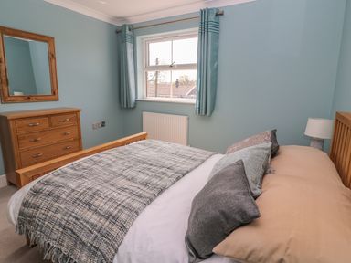 A bedroom with a wooden bed and a plaid blanket next to a wooden dresser with a mirror and a window with blue curtains at Cadwgan Aberporth
