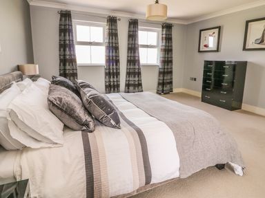 A bedroom with a bed with pillows and blanket two windows with curtains a black dresser and framed pictures on the wall at Cadwgan in Aberporth